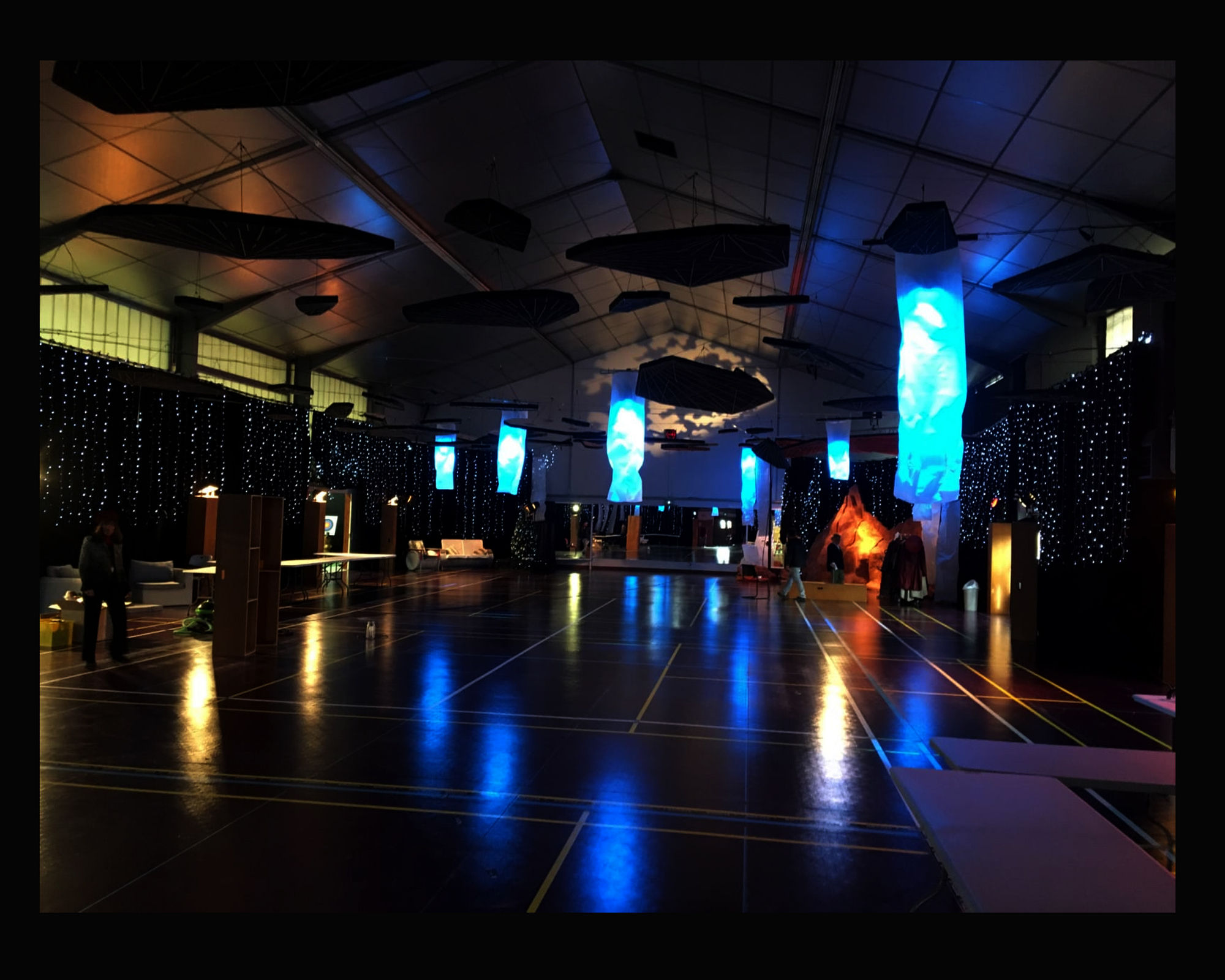 Décoration de salle pour un arbre de Noël sur le Thème de l'espace Transformation d'un gymnase, de 600 m2 : Pendrillonnage des murs, fabrication de tubes en papier calque éclairés de l'intérieur par des projecteurs LED, guirlandes lumineuses, suspension de plaques qui folttent dans la nuit 